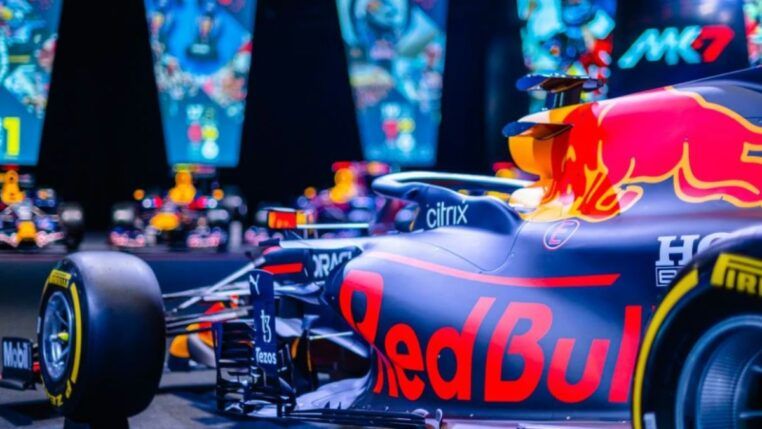 Carro de Fórmula 1 da Red Bull em destaque, com pintura azul e detalhes em vermelho e amarelo, exibido em ambiente iluminado com telas coloridas ao fundo mostrando imagens relacionadas à equipe.