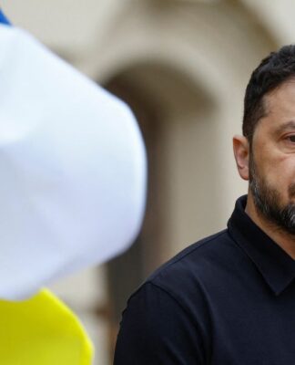 Homem de expressão séria, usando camisa preta, aparece em primeiro plano ao ar livre; à esquerda, uma bandeira azul e amarela desfocada ocupa parte da imagem.