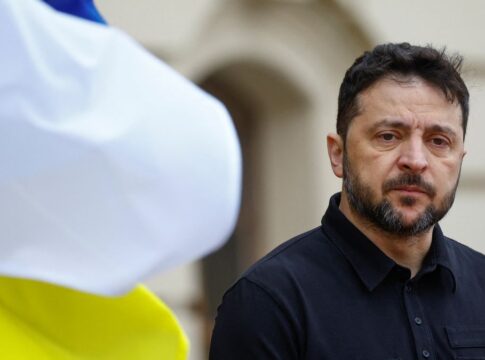 Homem de expressão séria, usando camisa preta, aparece em primeiro plano ao ar livre; à esquerda, uma bandeira azul e amarela desfocada ocupa parte da imagem.