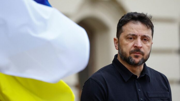 Homem de expressão séria, usando camisa preta, aparece em primeiro plano ao ar livre; à esquerda, uma bandeira azul e amarela desfocada ocupa parte da imagem.