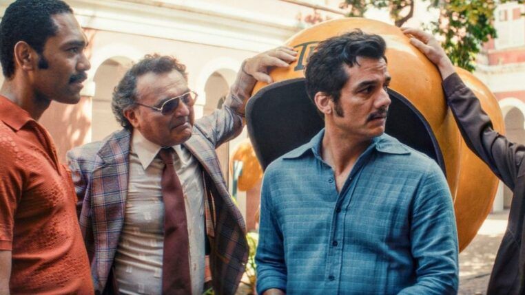 Imagem mostra uma cena do filme "O Agente Secreto": quatro homens em um pátio ensolarado em volta de um orelhão laranja. Na direita, estã o protagonista Wagner Moura