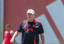 A imagem é um plano médio do técnico de futebol Abel Braga, em pé, em um campo de treinamento durante o dia, sob forte luz solar.