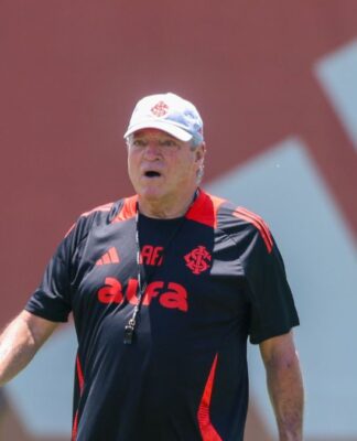 A imagem é um plano médio do técnico de futebol Abel Braga, em pé, em um campo de treinamento durante o dia, sob forte luz solar.
