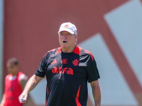 A imagem é um plano médio do técnico de futebol Abel Braga, em pé, em um campo de treinamento durante o dia, sob forte luz solar.