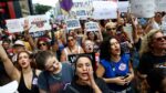 A imagem captura um momento vibrante de um protesto de rua composto majoritariamente por mulheres, carregando cartazes e manifestando-se com forte expressão emocional, provavelmente na Avenida Paulista, devido à densidade e ambiente urbano. O protesto é claramente contra o feminicídio e a misoginia.