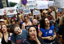 A imagem captura um momento vibrante de um protesto de rua composto majoritariamente por mulheres, carregando cartazes e manifestando-se com forte expressão emocional, provavelmente na Avenida Paulista, devido à densidade e ambiente urbano. O protesto é claramente contra o feminicídio e a misoginia.