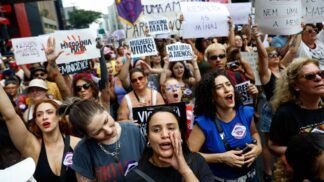 A imagem captura um momento vibrante de um protesto de rua composto majoritariamente por mulheres, carregando cartazes e manifestando-se com forte expressão emocional, provavelmente na Avenida Paulista, devido à densidade e ambiente urbano. O protesto é claramente contra o feminicídio e a misoginia.