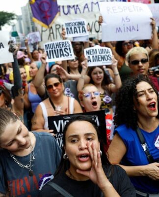 A imagem captura um momento vibrante de um protesto de rua composto majoritariamente por mulheres, carregando cartazes e manifestando-se com forte expressão emocional, provavelmente na Avenida Paulista, devido à densidade e ambiente urbano. O protesto é claramente contra o feminicídio e a misoginia.