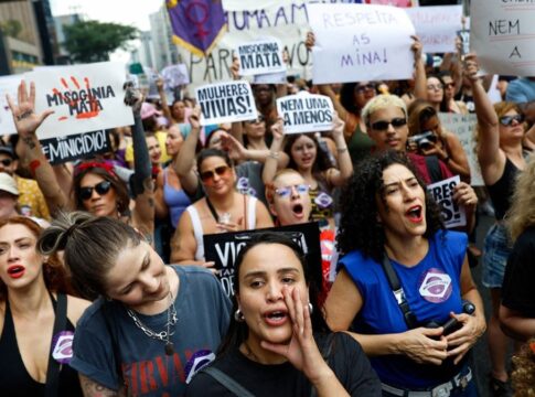 A imagem captura um momento vibrante de um protesto de rua composto majoritariamente por mulheres, carregando cartazes e manifestando-se com forte expressão emocional, provavelmente na Avenida Paulista, devido à densidade e ambiente urbano. O protesto é claramente contra o feminicídio e a misoginia.