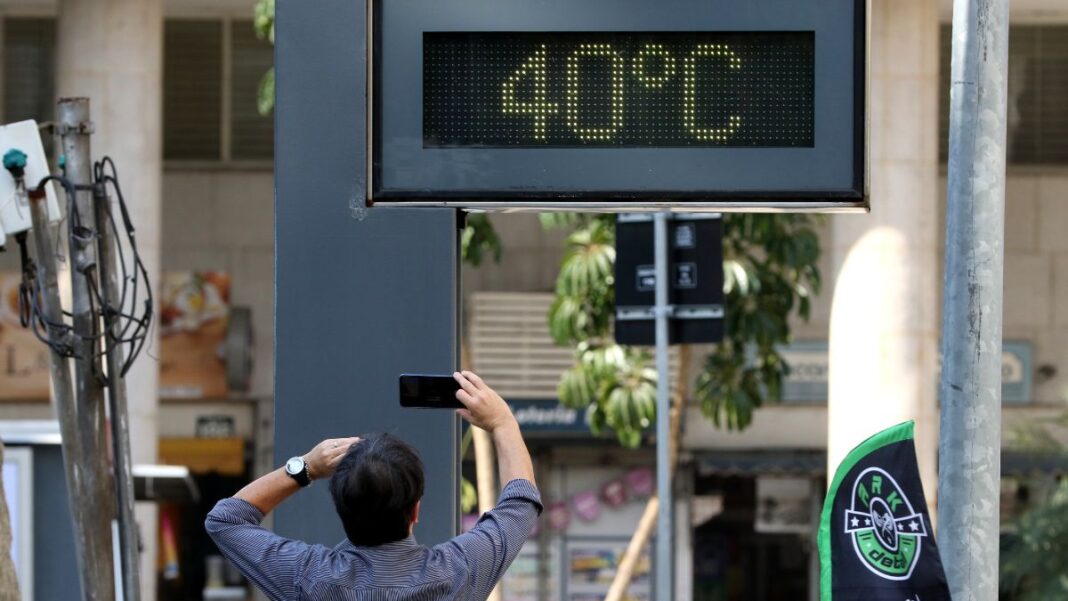 A foto registra um momento típico de calor extremo em um ambiente urbano. O foco central é um termômetro de rua digital que marca uma temperatura de 40ºC, enquanto um pedestre registra a cena.
