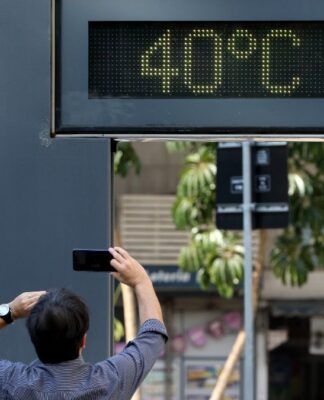 A foto registra um momento típico de calor extremo em um ambiente urbano. O foco central é um termômetro de rua digital que marca uma temperatura de 40ºC, enquanto um pedestre registra a cena.