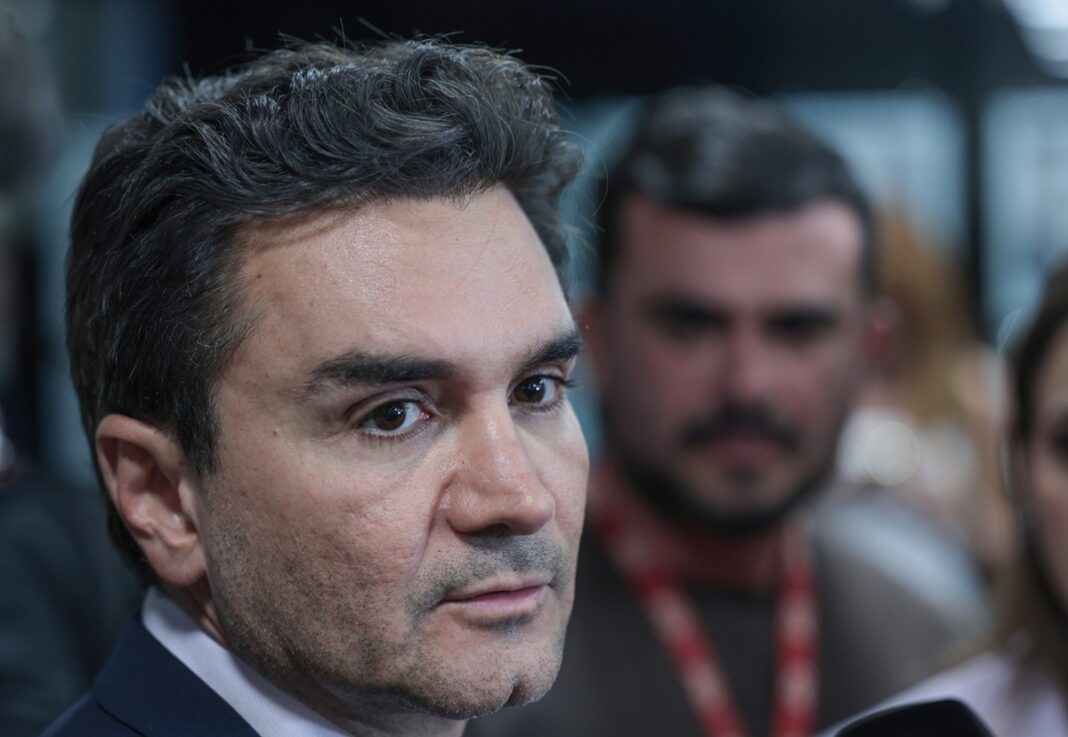 A foto é um primeiro plano (close-up) do rosto de Celso Sabino, atual Ministro do Turismo do Brasil. Ele está posicionado de três quartos, olhando para a direita da imagem com uma expressão séria e atenta, como se estivesse ouvindo uma pergunta ou concedendo uma entrevista.