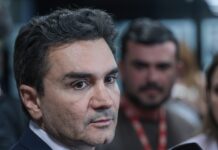 A foto é um primeiro plano (close-up) do rosto de Celso Sabino, atual Ministro do Turismo do Brasil. Ele está posicionado de três quartos, olhando para a direita da imagem com uma expressão séria e atenta, como se estivesse ouvindo uma pergunta ou concedendo uma entrevista.