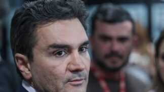 A foto é um primeiro plano (close-up) do rosto de Celso Sabino, atual Ministro do Turismo do Brasil. Ele está posicionado de três quartos, olhando para a direita da imagem com uma expressão séria e atenta, como se estivesse ouvindo uma pergunta ou concedendo uma entrevista.