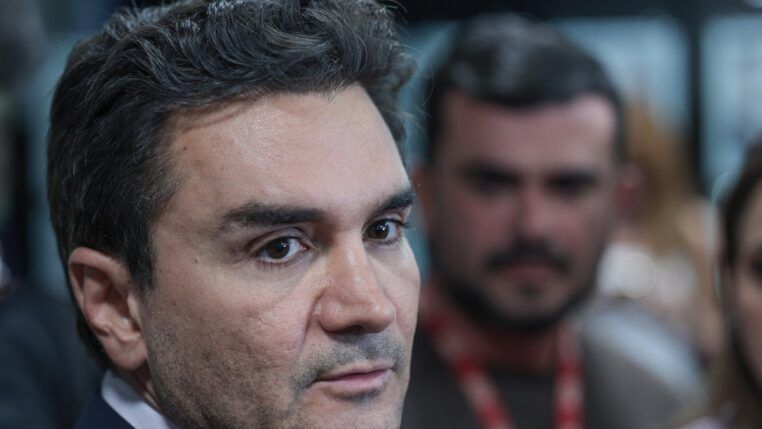 A foto é um primeiro plano (close-up) do rosto de Celso Sabino, atual Ministro do Turismo do Brasil. Ele está posicionado de três quartos, olhando para a direita da imagem com uma expressão séria e atenta, como se estivesse ouvindo uma pergunta ou concedendo uma entrevista.