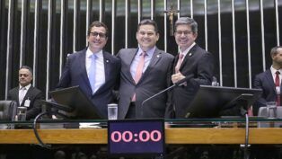 A imagem captura três parlamentares no centro da mesa principal de um plenário do Congresso Nacional, interagindo em um momento de aparente celebração ou satisfação, possivelmente após a aprovação da Lei de Diretrizes Orçamentárias (LDO), já que o relator da LDO (Gervásio Maia) e o presidente do Congresso (Davi Alcolumbre) estão na foto.