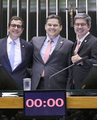 A imagem captura três parlamentares no centro da mesa principal de um plenário do Congresso Nacional, interagindo em um momento de aparente celebração ou satisfação, possivelmente após a aprovação da Lei de Diretrizes Orçamentárias (LDO), já que o relator da LDO (Gervásio Maia) e o presidente do Congresso (Davi Alcolumbre) estão na foto.