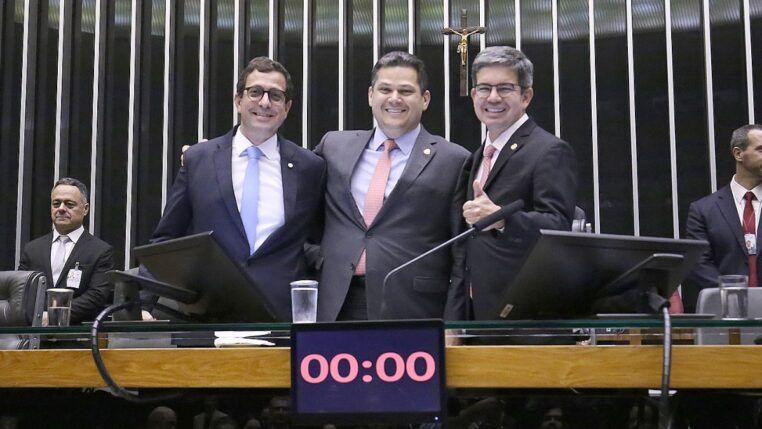 A imagem captura três parlamentares no centro da mesa principal de um plenário do Congresso Nacional, interagindo em um momento de aparente celebração ou satisfação, possivelmente após a aprovação da Lei de Diretrizes Orçamentárias (LDO), já que o relator da LDO (Gervásio Maia) e o presidente do Congresso (Davi Alcolumbre) estão na foto.