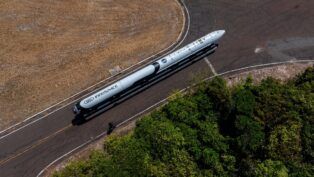 Esta é uma foto aérea (tirada de cima) que mostra um foguete sendo transportado por uma estrada ou pista em uma área predominantemente rural ou de vegetação.