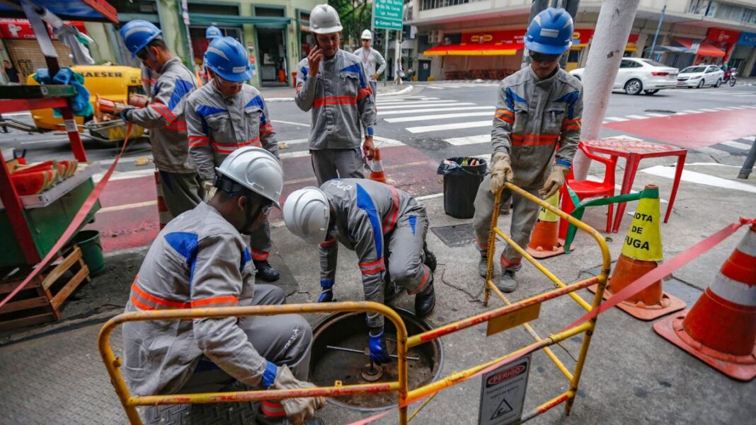A imagem mostra um grupo de cerca de seis trabalhadores da empresa de energia Enel realizando manutenção ou reparos em uma via urbana de São Paulo. Eles estão em uma pose de trabalho concentrado ao redor de um bueiro ou abertura no chão.