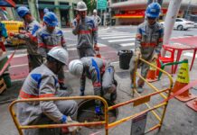 A imagem mostra um grupo de cerca de seis trabalhadores da empresa de energia Enel realizando manutenção ou reparos em uma via urbana de São Paulo. Eles estão em uma pose de trabalho concentrado ao redor de um bueiro ou abertura no chão.