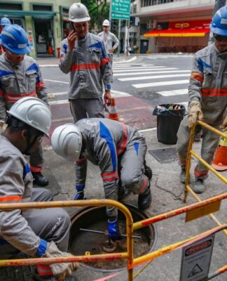 A imagem mostra um grupo de cerca de seis trabalhadores da empresa de energia Enel realizando manutenção ou reparos em uma via urbana de São Paulo. Eles estão em uma pose de trabalho concentrado ao redor de um bueiro ou abertura no chão.