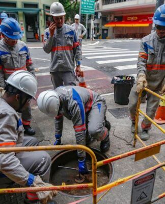 A imagem mostra um grupo de cerca de seis trabalhadores da empresa de energia Enel realizando manutenção ou reparos em uma via urbana de São Paulo. Eles estão em uma pose de trabalho concentrado ao redor de um bueiro ou abertura no chão.
