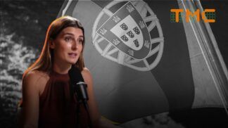 A imagem mostra a jornalista Joana Treptow em um plano médio, sentada e falando em um microfone de estúdio, com uma bandeira de Portugal em preto e branco ao fundo.