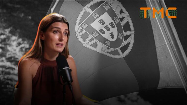 A imagem mostra a jornalista Joana Treptow em um plano médio, sentada e falando em um microfone de estúdio, com uma bandeira de Portugal em preto e branco ao fundo.