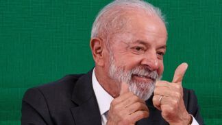 A foto mostra um plano médio do presidente Luiz Inácio Lula da Silva, que está sorrindo e gesticulando com as duas mãos em um sinal de aprovação ("joinha" ou polegar para cima).