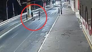 A imagem é um registro de câmera de monitoramento (CFTV), com ângulo alto e diagonal, mostrando uma via pública urbana durante a noite ou madrugada. A iluminação é artificial e fria, vindo de postes de luz. No meio da rua, destacado por um grande círculo vermelho desenhado digitalmente na imagem, há um grupo de três pessoas em pé ao redor de uma quarta figura que parece estar caída ou sentada no chão.