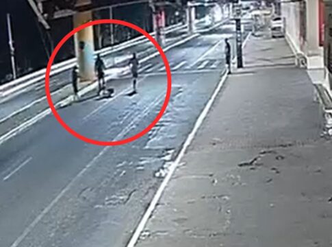 A imagem é um registro de câmera de monitoramento (CFTV), com ângulo alto e diagonal, mostrando uma via pública urbana durante a noite ou madrugada. A iluminação é artificial e fria, vindo de postes de luz. No meio da rua, destacado por um grande círculo vermelho desenhado digitalmente na imagem, há um grupo de três pessoas em pé ao redor de uma quarta figura que parece estar caída ou sentada no chão.