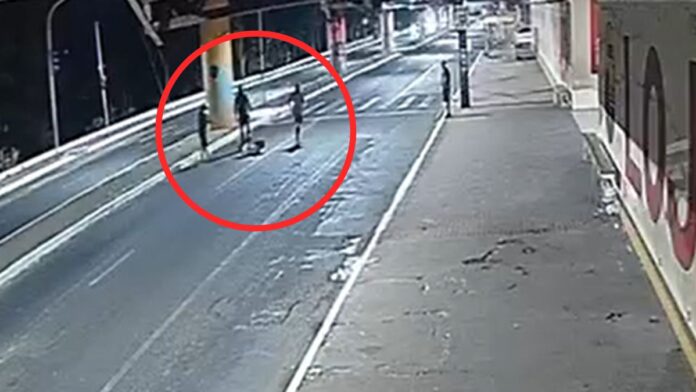 A imagem é um registro de câmera de monitoramento (CFTV), com ângulo alto e diagonal, mostrando uma via pública urbana durante a noite ou madrugada. A iluminação é artificial e fria, vindo de postes de luz. No meio da rua, destacado por um grande círculo vermelho desenhado digitalmente na imagem, há um grupo de três pessoas em pé ao redor de uma quarta figura que parece estar caída ou sentada no chão.