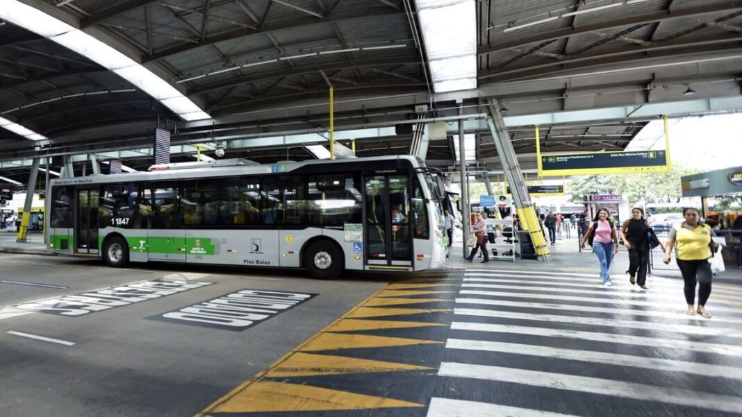 A imagem mostra um terminal de ônibus coberto na cidade de São Paulo, com um veículo de transporte público parado e pessoas circulando na área de embarque e desembarque.