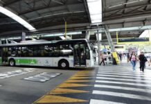 A imagem mostra um terminal de ônibus coberto na cidade de São Paulo, com um veículo de transporte público parado e pessoas circulando na área de embarque e desembarque.