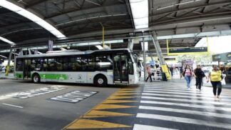 A imagem mostra um terminal de ônibus coberto na cidade de São Paulo, com um veículo de transporte público parado e pessoas circulando na área de embarque e desembarque.