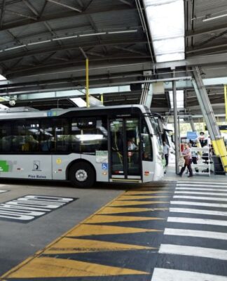 A imagem mostra um terminal de ônibus coberto na cidade de São Paulo, com um veículo de transporte público parado e pessoas circulando na área de embarque e desembarque.