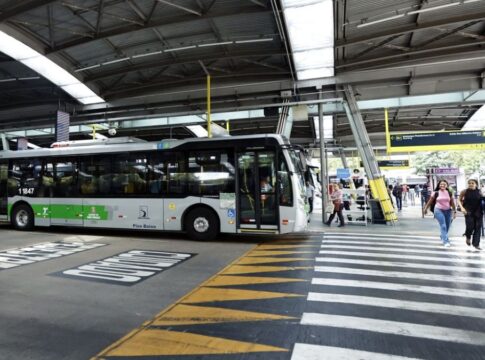A imagem mostra um terminal de ônibus coberto na cidade de São Paulo, com um veículo de transporte público parado e pessoas circulando na área de embarque e desembarque.