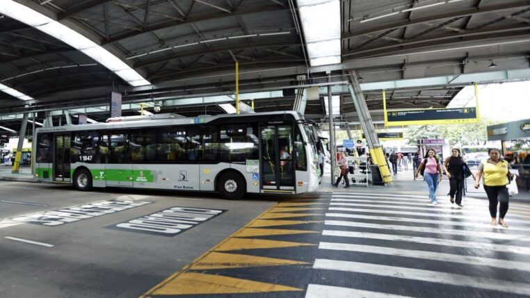 A imagem mostra um terminal de ônibus coberto na cidade de São Paulo, com um veículo de transporte público parado e pessoas circulando na área de embarque e desembarque.