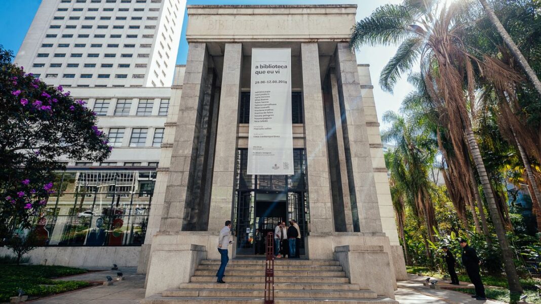 A imagem é uma vista ampla e frontal do edifício principal da Biblioteca Mário de Andrade, mostrando sua arquitetura imponente e o entorno verde, sob luz solar intensa. O prédio é construído em um estilo monumental e clássico, com predomínio de pedra clara ou granito cinza nas fachadas. A entrada principal é marcada por uma grande escadaria que leva a uma plataforma elevada.