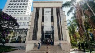 A imagem é uma vista ampla e frontal do edifício principal da Biblioteca Mário de Andrade, mostrando sua arquitetura imponente e o entorno verde, sob luz solar intensa. O prédio é construído em um estilo monumental e clássico, com predomínio de pedra clara ou granito cinza nas fachadas. A entrada principal é marcada por uma grande escadaria que leva a uma plataforma elevada.