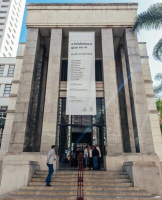 A imagem é uma vista ampla e frontal do edifício principal da Biblioteca Mário de Andrade, mostrando sua arquitetura imponente e o entorno verde, sob luz solar intensa. O prédio é construído em um estilo monumental e clássico, com predomínio de pedra clara ou granito cinza nas fachadas. A entrada principal é marcada por uma grande escadaria que leva a uma plataforma elevada.