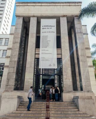 A imagem é uma vista ampla e frontal do edifício principal da Biblioteca Mário de Andrade, mostrando sua arquitetura imponente e o entorno verde, sob luz solar intensa. O prédio é construído em um estilo monumental e clássico, com predomínio de pedra clara ou granito cinza nas fachadas. A entrada principal é marcada por uma grande escadaria que leva a uma plataforma elevada.