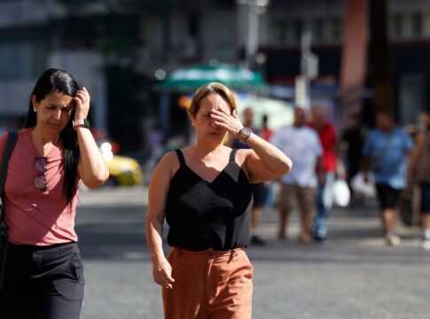 A foto captura um momento cotidiano de pedestres caminhando sob sol forte em um centro urbano. O foco principal está em duas mulheres em primeiro plano que demonstram desconforto com a luminosidade e a temperatura elevada.