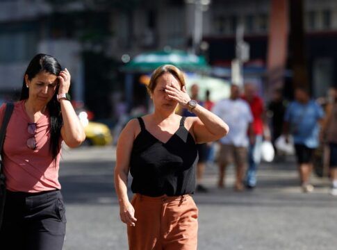 A foto captura um momento cotidiano de pedestres caminhando sob sol forte em um centro urbano. O foco principal está em duas mulheres em primeiro plano que demonstram desconforto com a luminosidade e a temperatura elevada.