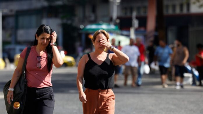 A foto captura um momento cotidiano de pedestres caminhando sob sol forte em um centro urbano. O foco principal está em duas mulheres em primeiro plano que demonstram desconforto com a luminosidade e a temperatura elevada.