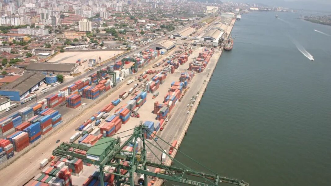 Esta é uma vista aérea ampla que captura uma seção do Porto de Santos no Brasil, destacando a intensa atividade de movimentação de contêineres e a proximidade com a área urbana da cidade.