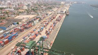 Esta é uma vista aérea ampla que captura uma seção do Porto de Santos no Brasil, destacando a intensa atividade de movimentação de contêineres e a proximidade com a área urbana da cidade.