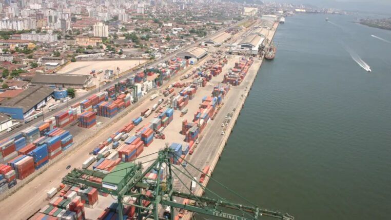 Esta é uma vista aérea ampla que captura uma seção do Porto de Santos no Brasil, destacando a intensa atividade de movimentação de contêineres e a proximidade com a área urbana da cidade.