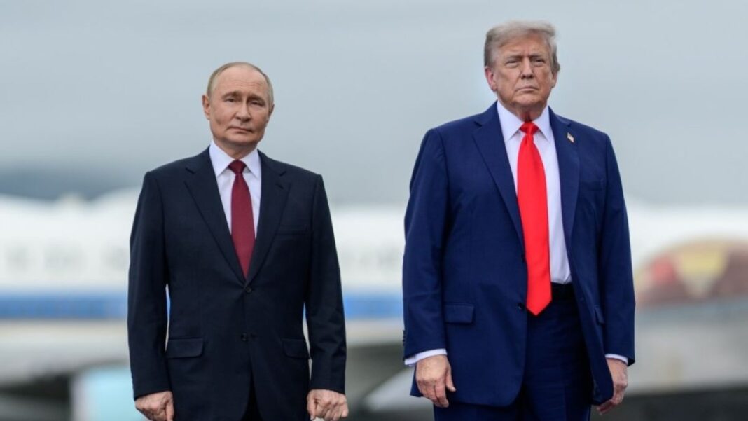 A imagem é um plano médio que captura os presidentes Vladimir Putin e Donald Trump lado a lado, em pé, em um ambiente externo que se assemelha a uma pista de aeroporto ou área externa de recepção, sob um céu nublado.